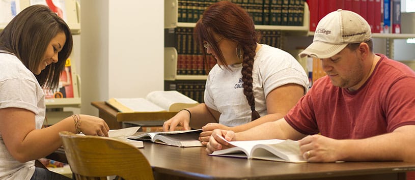A group of three students study at a large table with a variety of textbooks
