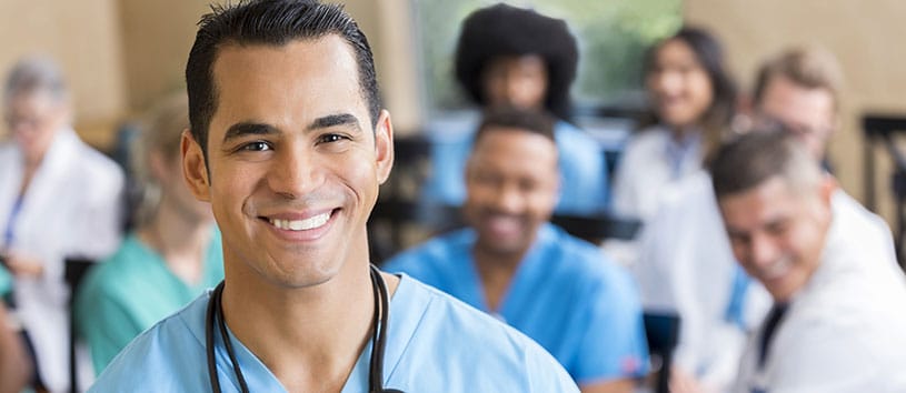 Male nurse standing in a healthcare environment with medical colleagues behind him.